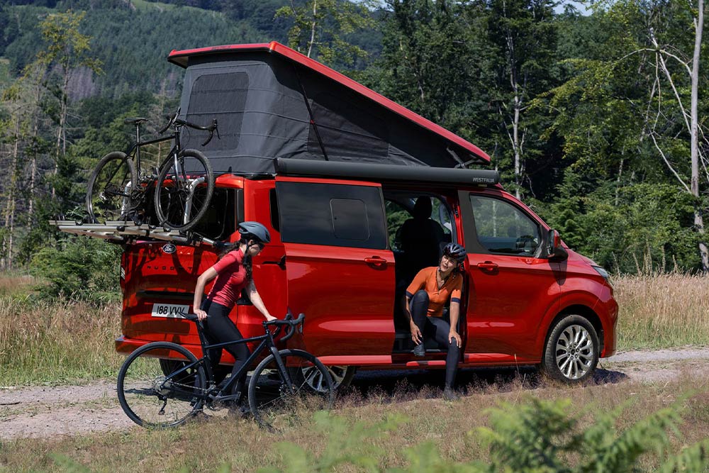 Des personnes déchargent des vélos du porte-vélos d’un Ford Nugget rouge.