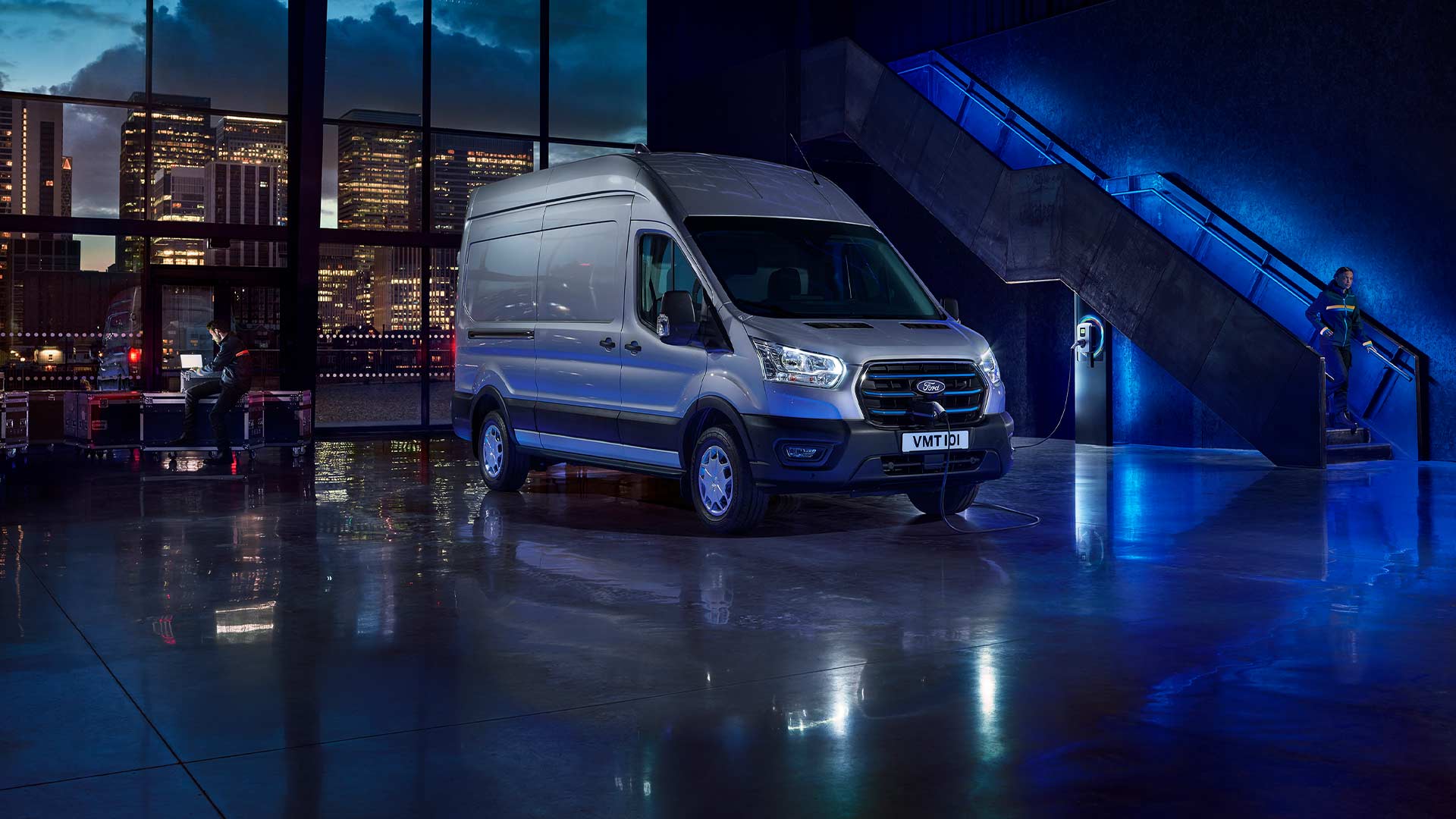 Fourgon électrique Ford E-Transit argenté garé à l'intérieur avec un paysage urbain visible à travers de grandes fenêtres. 