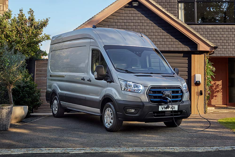 Fourgon électrique Ford E-Transit argenté garé devant dans une allée à côté d'une maison et connecté à une borne de recharge.