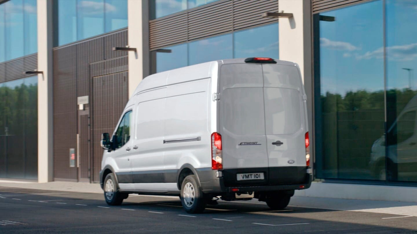 Ford E-Transit, couleur argent, vue arrière aux trois quarts, stationnant devant un bâtiment moderne
