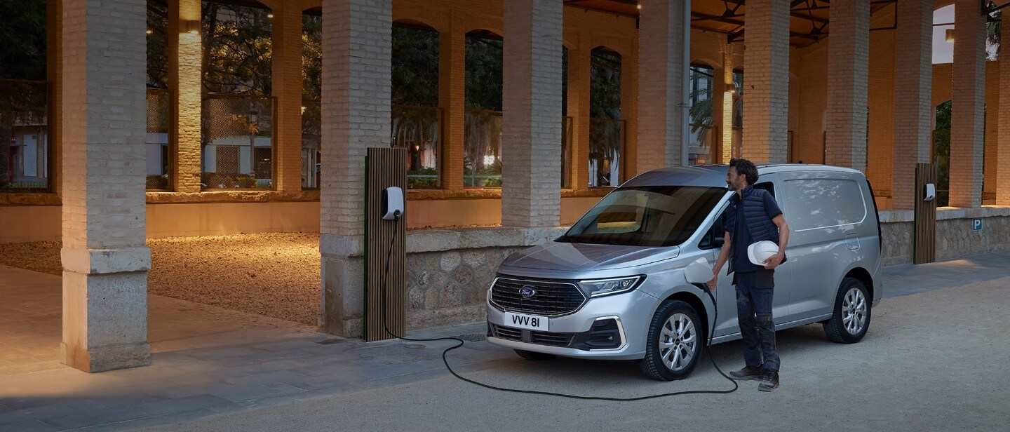 Un homme équipé d’un casque de protection branche un câble de recharge Wallbox sur un Ford Transit Connect de couleur Stardust Silver.
