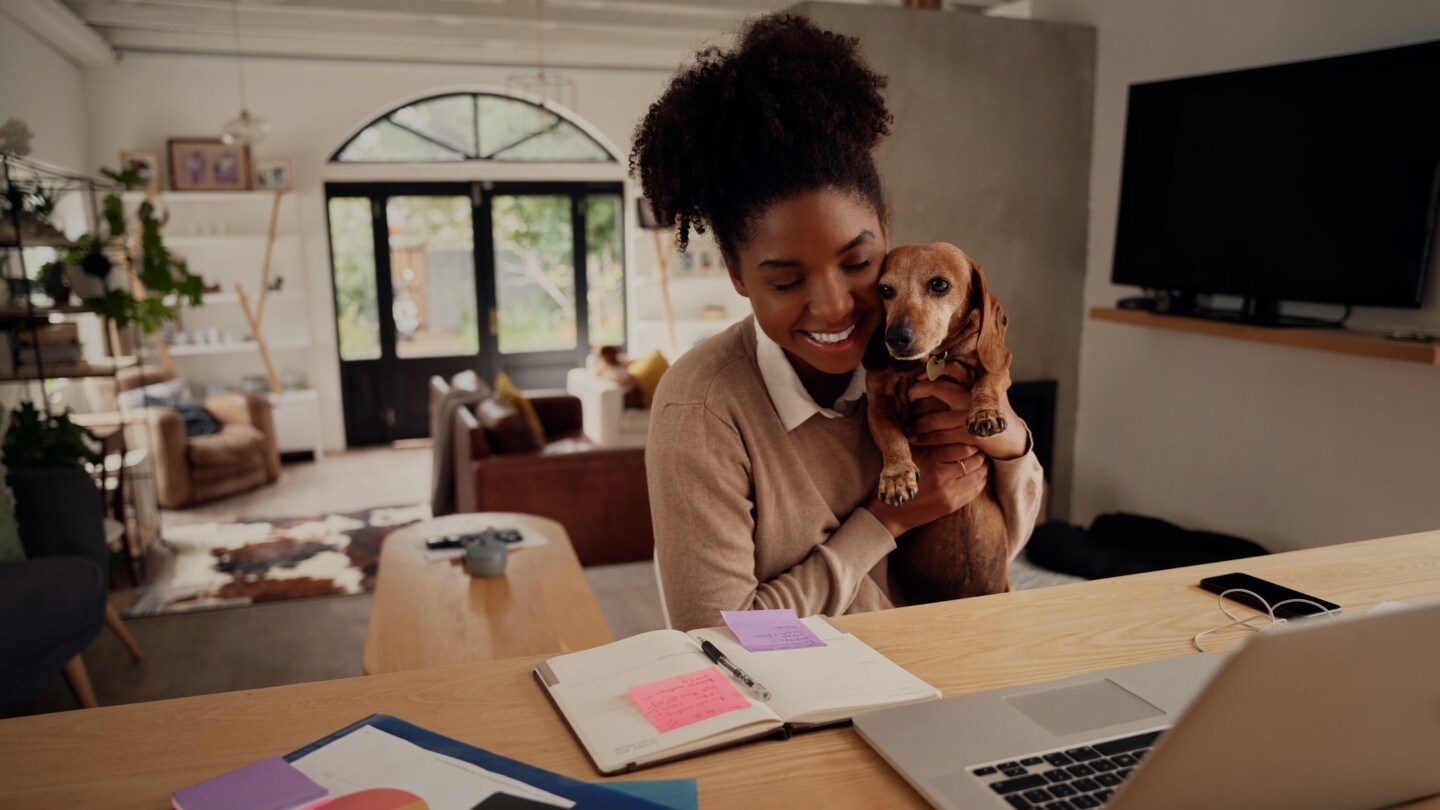 Femme souriante dans sa maison avec un chien dans les bras, devant un ordinateur portable