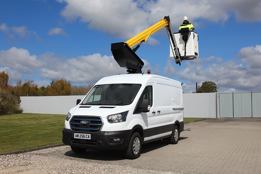 Une camionnette Ford Transit blanche avec une nacelle élévatrice étendue, un technicien dans le panier, sous un ciel bleu.