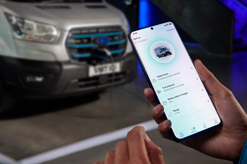 Une personne tenant un smartphone affichant une interface d'application, avec une camionnette Ford Pro Transit argentée floue en arrière-plan.