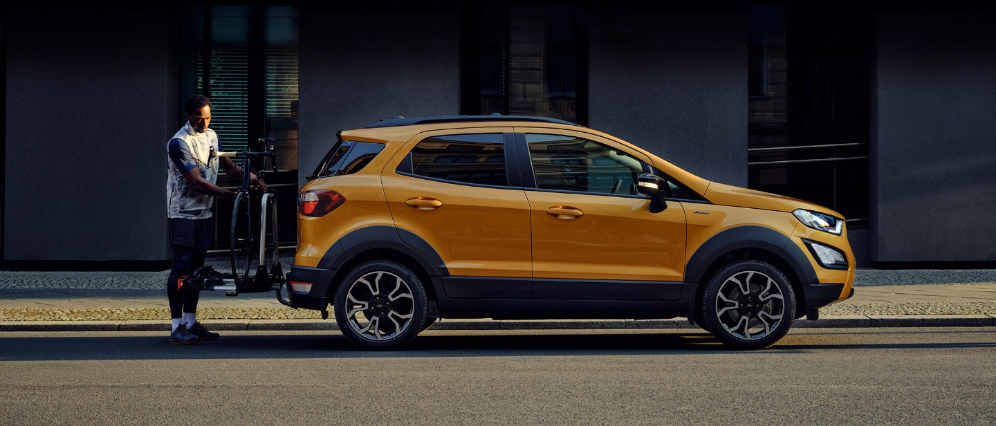 Vue latérale du Ford EcoSport couleur orange, parqué dans une rue en ville pendant qu’un homme fixe un vélo sur le porte-vélos arrière. Vue latérale du Ford EcoSport couleur orange, parqué dans une rue en ville pendant qu’un homme fixe un vélo sur le porte-vélos arrière.