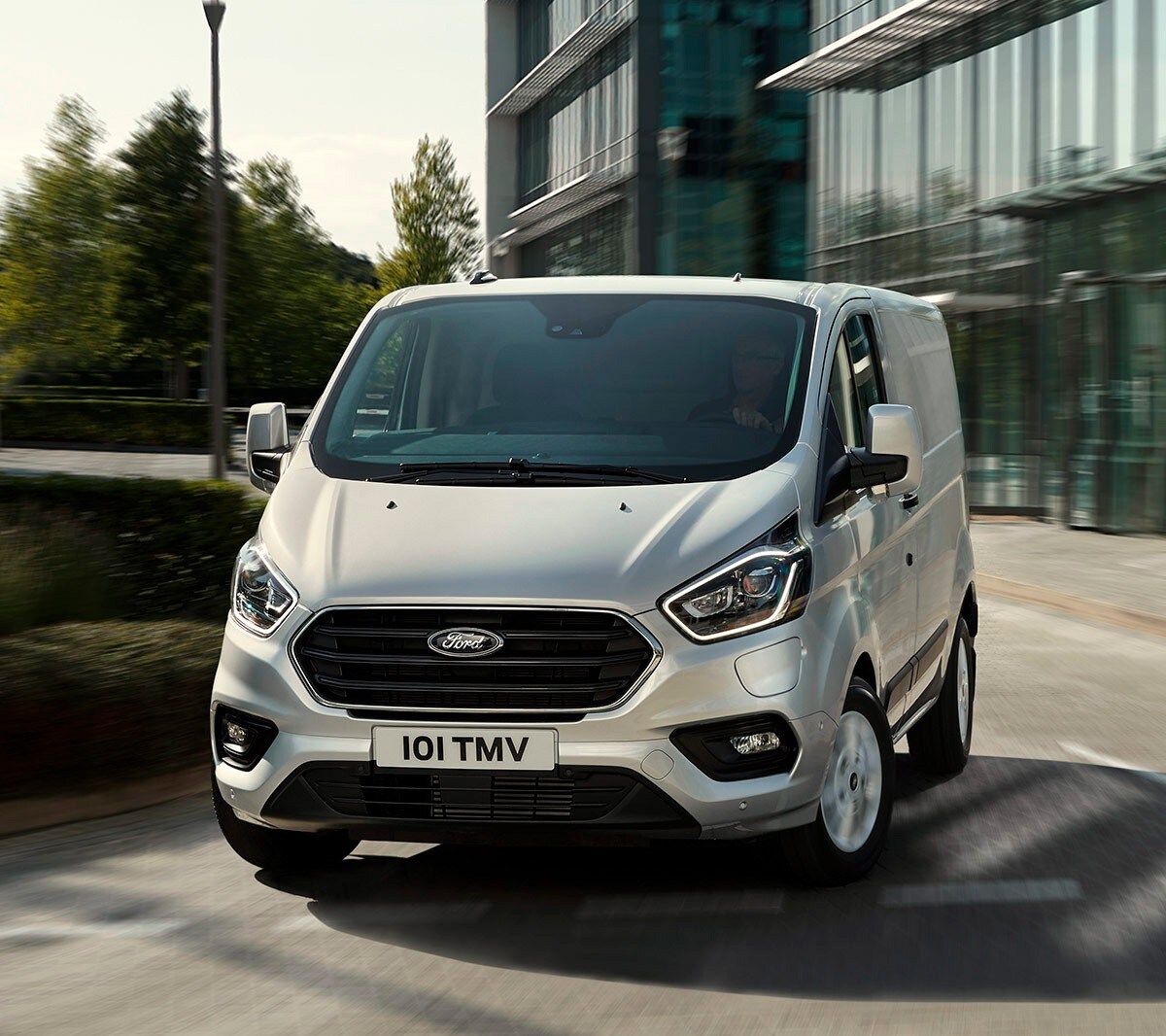 Ford Transit Custom argenté roulant devant un bâtiment avec une façade en verre