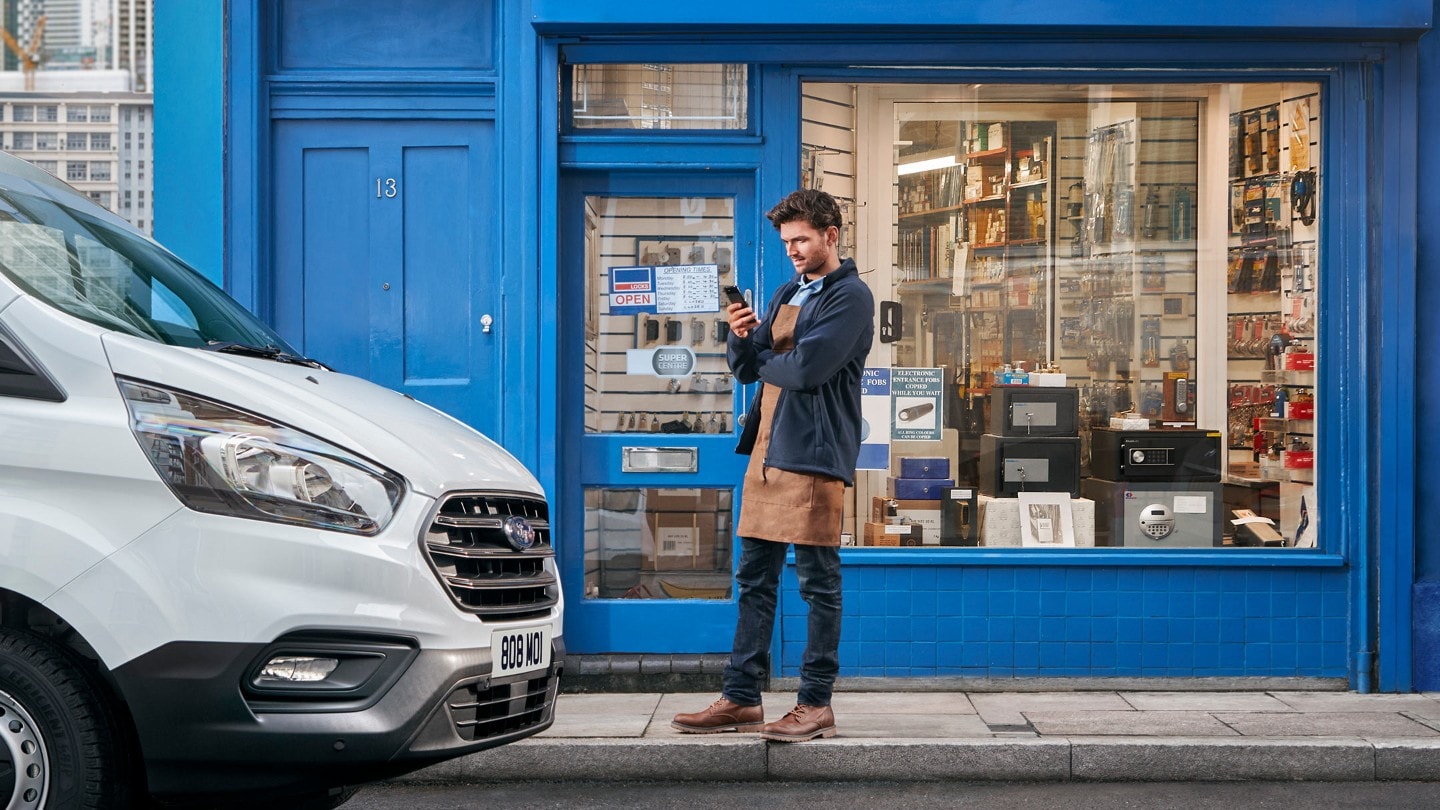 Un homme se tient devant un magasin à côté d'un véhicule d'entreprise, un téléphone portable à la main.