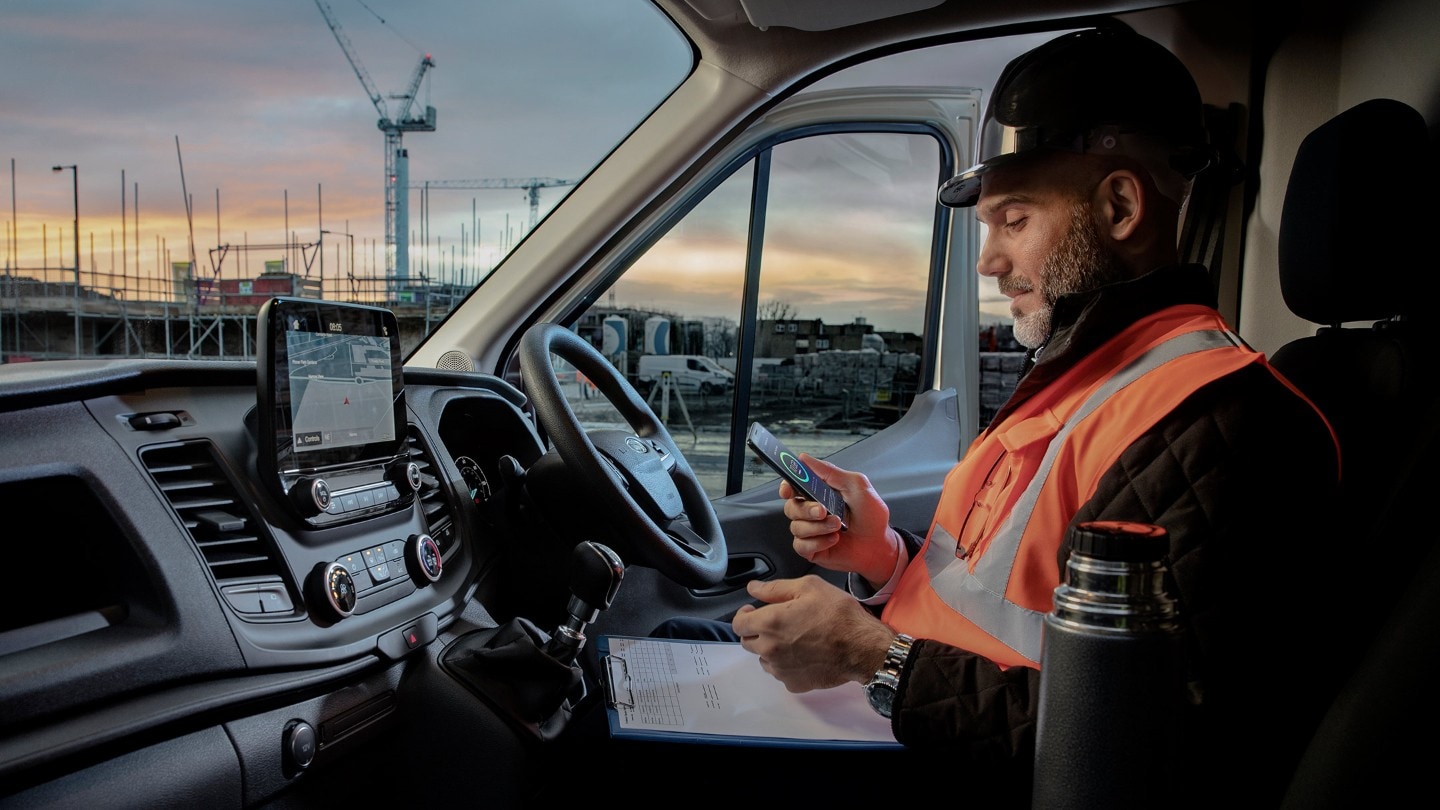 Ouvrier portant un casque de chantier assis derrière le volant d'un véhicule et regardant un téléphone portable 
