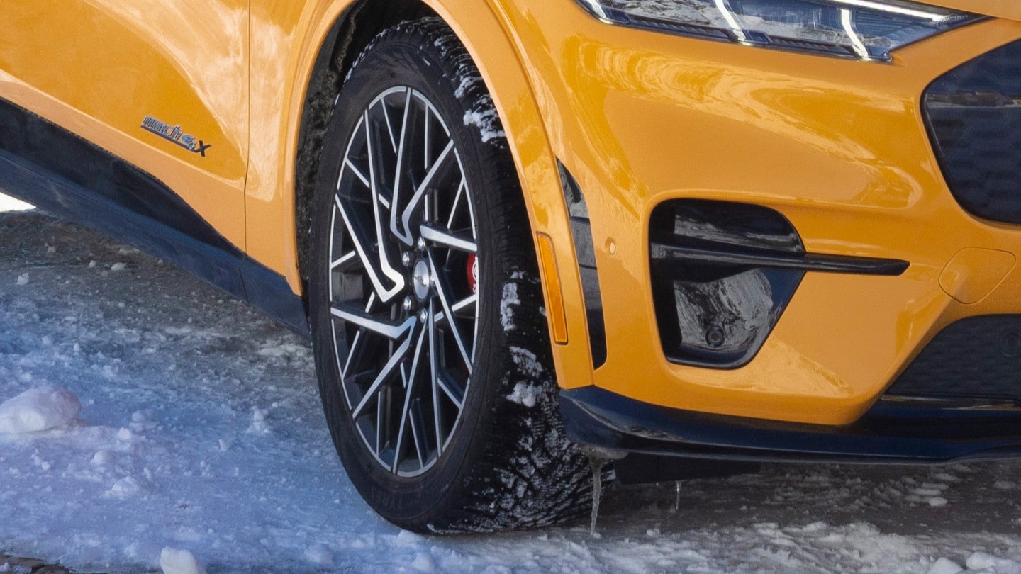 Gros plan sur la roue avant et l’aile d’un véhicule jaune vif – vraisemblablement une Ford Mustang Mach-E – parqué sur une surface enneigée.