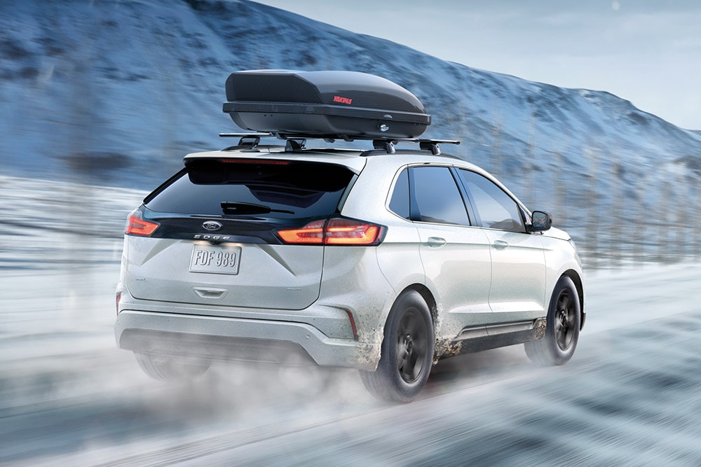 Une Ford Edge blanche avec un coffre de toit roulant sur une route enneigée, vue de trois quarts arrière.
